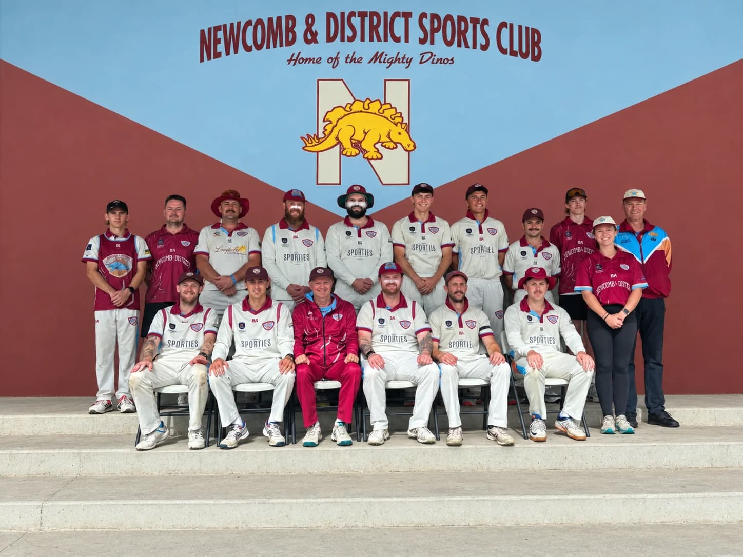 NDCC GCA Division 4 1st XI premiership team: players and officials in maroon and white club kit, posed in two rows, standing and seated, in front of the Newcomb & District Sports Club "Home of the Mighty Dinos" mural.